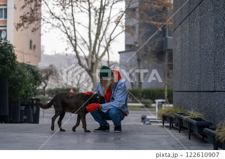 Kind, warm-hearted stylish guy in trendy outfit sitting on haunches, caressing hungry street dog 122610907