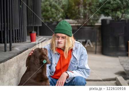 Caring young guy in stylish clothes strokes a homeless microchipped dog on the street on summer day in the park. Concept of caring for abandoned animals. Volunteer man at a shelter for homeless pets 122610916