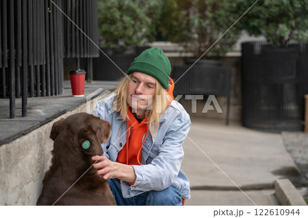 Kind warm-hearted stylish guy in trendy outfit sitting on haunches, caressing street dog with love Kind warm-hearted stylish guy in trendy outfit sitting on haunches, caressing street dog with love 122610944