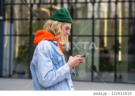 Pleased hipster guy standing outside with smartphone, looking screen typing message in social media. 122611010