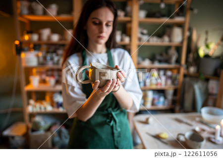 Female artisan examining freshly made clay cup, evaluating form and texture in pottery studio class 122611056