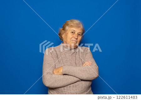Caucasian woman about 70s wearing dark beige sweater shrugging shoulders, feeling confused and unsure, doubting with arms crossed. Isolated over blue background 122611483