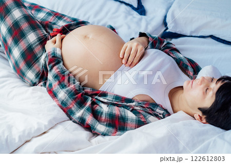 Pregnant woman lying on bed, touching her belly, showcasing joy and anticipation. Expecting mother relaxing in pajama during last trimester of pregnancy. Motherhood, preparation and expectation. Pregnant woman lying on bed, touching her belly, showcasing joy and anticipation. Expecting mother relaxing in pajama during last trimester of pregnancy. Motherhood, preparation and expectation. 122612803