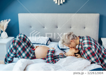 Mixed race couple are waiting for a baby. Husband and his pregnant wife in cozy pajamas relaxing on the bed at modern home bedroom. Romantic moments for pregnant couple, warm atmosphere of love. 122612804