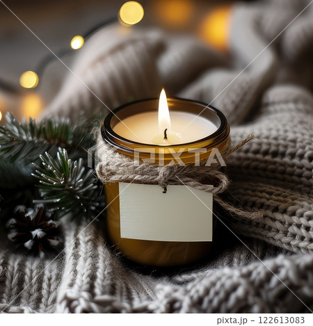 Scented candle in glass jar on soft grey jersey fabric, Winter product photo. Warm glow of scented candle creates cozy winter atmosphere. Scented candle jar with blank label mockup 122613083
