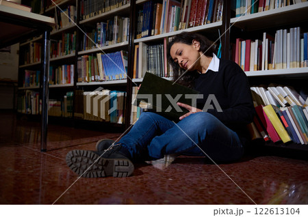 Young woman reading a book while sitting on the library floor 122613104