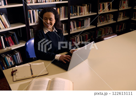 Smiling woman using a laptop in a library for studying or working Smiling woman using a laptop in a library for studying or working 122613111