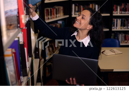 Smiling woman reaching for book in library holding a laptop Smiling woman reaching for book in library holding a laptop 122613125
