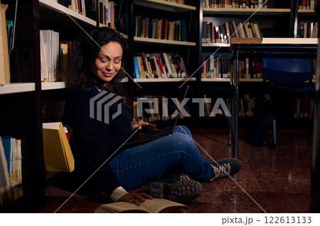 Young woman studying with a book and laptop in a cozy library corner Young woman studying with a book and laptop in a cozy library corner 122613133