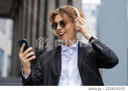 Happy joyful businessman listens to music on wireless headphones while walking on street near office 122614103