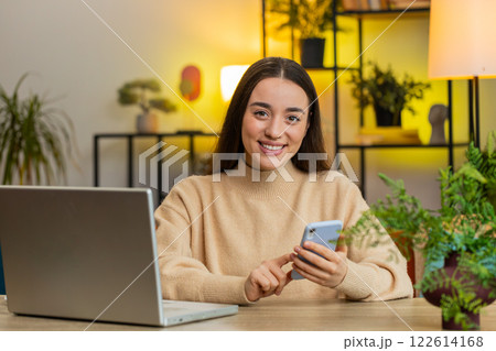 Caucasian young woman sits at home table use smartphone share messages on social media application 122614168