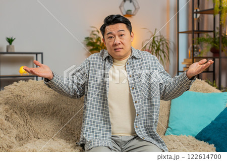 Confused Asian handsome man feeling embarrassed about ambiguous question sitting on sofa at home Confused Asian handsome man feeling embarrassed about ambiguous question sitting on sofa at home 122614700