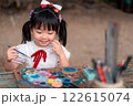 Smiling Asian Girl Engaged in Creative Art Activity with Colorful Paints and Brushes Outdoors 122615074
