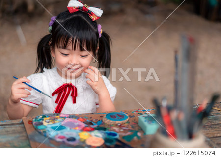 Smiling Asian Girl Engaged in Creative Art Activity with Colorful Paints and Brushes Outdoors 122615074