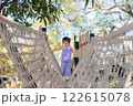 Young Asian Girl Playing on Rope Bridge in Natural Outdoor Setting with Greenery and Bright Sky 122615078