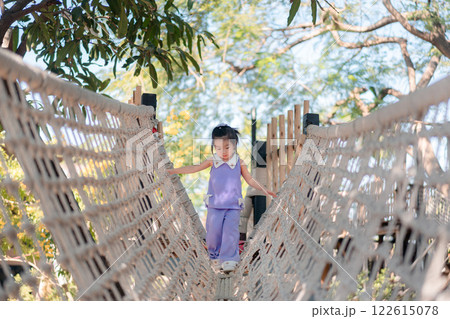 Young Asian Girl Playing on Rope Bridge in Natural Outdoor Setting with Greenery and Bright Sky Young Asian Girl Playing on Rope Bridge in Natural Outdoor Setting with Greenery and Bright Sky 122615078
