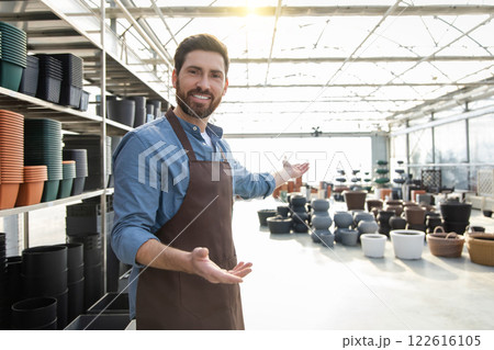 Man worker in garden center wearing uniform, arranging many pots for plants on racks and shelves 122616105