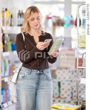 Positive happy young girl buyer in shop holding and choosing dishwashing liquid, scan QR code 122617735