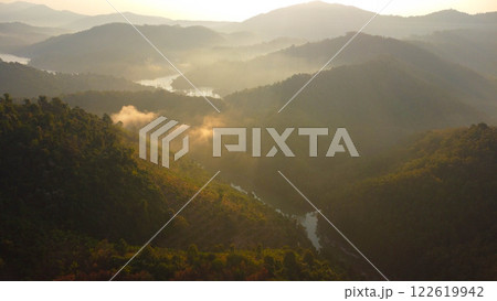 Beautiful sunrise on the mountain in winter. The sun shines down on a serene mountain landscape covered with tropical trees and the calm river in the early morning. 122619942