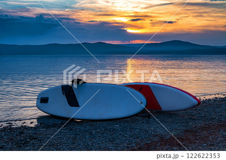Two sup boards on the shore of a picturesque lake against the backdrop of sunset 122622353