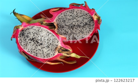 Organic dragon fruit sliced open reveals speckled white flesh with black seeds placed on vibrant red plate against blue background. Eco presentation highlights organic freshness and vibrant colors. 122623217