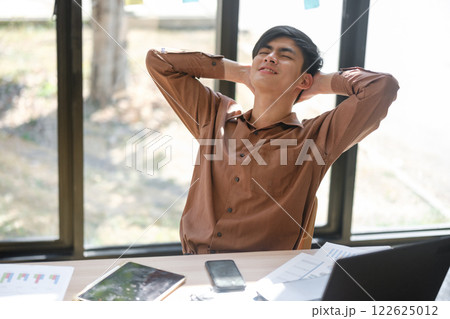 Young man stretching and leaning back to relax after long hours of work at his desk 122625012