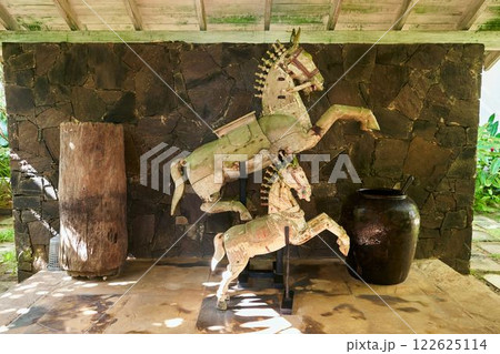 Vintage tropical villa interior with rustic wooden horse sculptures, large stone vase. Natural light highlights unique decor. Cozy space combines antique, tropical elements. Ideal for bohemian style. Vintage tropical villa interior with rustic wooden horse sculptures, large stone vase. Natural light highlights unique decor. Cozy space combines antique, tropical elements. Ideal for bohemian style. 122625114