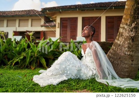 Gender fluid person in white wedding dress sits by palm tree, embracing tropical scenery. LGBTQ bridal concept. Calm moment at exotic destination. Modern identity, style in inclusive wedding fashion. 122625150