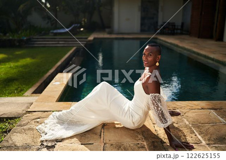 Black person in stylish white bridal dress poses by villa pool. Gender fluid fashion trend. LGBTQ friendly setting for chic wedding or event. Ethnic diversity. Summer day vibe. 122625155