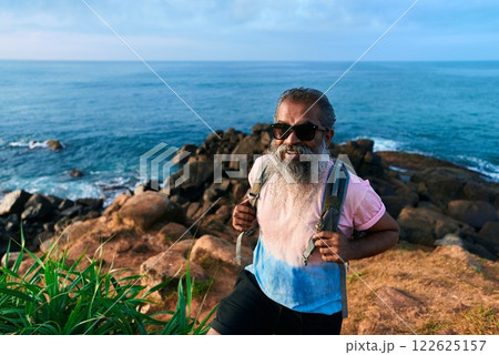 Senior ethnic man hikes on rocky coastal path, enjoys nature. Ocean view, backpack adventure. Elderly active lifestyle, explore outdoors. Sunny day, rugged terrain, vibrant sea backdrop. Senior ethnic man hikes on rocky coastal path, enjoys nature. Ocean view, backpack adventure. Elderly active lifestyle, explore outdoors. Sunny day, rugged terrain, vibrant sea backdrop. 122625157