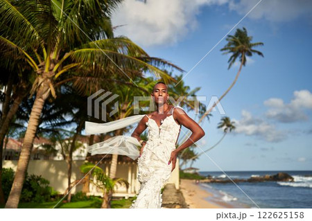 Person in white bridal dress on tropical beach. Ambiguous gender fluid black model poses between palm trees, ocean in background. LGBTQ-friendly wedding concept. Destination bridal fashion. 122625158