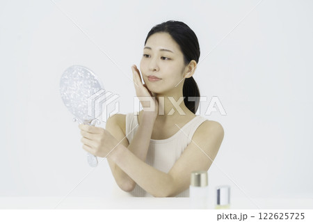 白バックのタンクトップを着て鏡を見ながら手を頬に当てる笑顔の若い女性　ビューティー 122625725