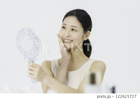 白バックのタンクトップを着て鏡を見ながら手を頬に当てる笑顔の若い女性 ビューティー 白バックのタンクトップを着て鏡を見ながら手を頬に当てる笑顔の若い女性 ビューティー 122625727