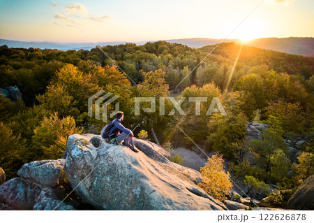 Aerial view of climber sitting atop rock formation at Dovbush Rocks in Carpathian mountains, Ukraine. Sun sets, casting warm glow over autumn forest and distant hills, creating breathtaking view. 122626878