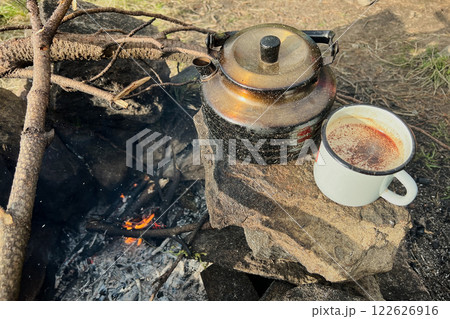 White enamel mug filled with hot beverage sits beside rustic black kettle on stone fire pit. Nearby, some sticks arranged for fire, all set against a backdrop of a green, forested camping area. White enamel mug filled with hot beverage sits beside rustic black kettle on stone fire pit. Nearby, some sticks arranged for fire, all set against a backdrop of a green, forested camping area. 122626916