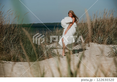 Woman wrapped in white blanket walking barefoot through sandy grasslands near the ocean on sunny day Woman wrapped in white blanket walking barefoot through sandy grasslands near the ocean on sunny day 122630320