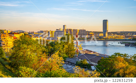 The Sava River flows peacefully past the Kalamegdan fortress as the sun sets, casting a warm glow on the cityscape of Belgrade. The view captures a vibrant blend of nature and urban life. 122631259