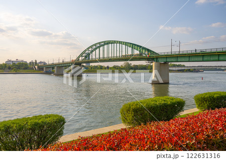 The Old Sava Bridge gracefully arches over the Sava River in Belgrade, showcasing its green structure against the clear sky, with lush greenery and vibrant flowers along the riverbank. The Old Sava Bridge gracefully arches over the Sava River in Belgrade, showcasing its green structure against the clear sky, with lush greenery and vibrant flowers along the riverbank. 122631316