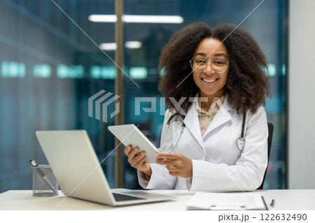 Smiling African American woman doctor using tablet in office. Wearing white coat and stethoscope, representing technology in healthcare. Promotes modern communication and patient engagement. 122632490