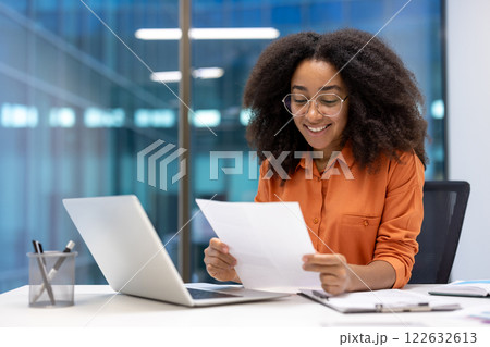Successful female financier holds a report document in her hands, reads it and smiles. Businesswoman is satisfied with the results of her work at the workplace inside the office. Successful female financier holds a report document in her hands, reads it and smiles. Businesswoman is satisfied with the results of her work at the workplace inside the office. 122632613