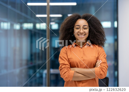 Confident young professional woman with glasses standing arms crossed, smiling warmly in modern office. Represents empowerment, success, and leadership in business setting. 122632620