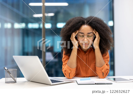 Young businesswoman sitting at desk, stressed while working on laptop. Hands holding head in frustration, illustrating challenges of workplace pressure and digital demands. Young businesswoman sitting at desk, stressed while working on laptop. Hands holding head in frustration, illustrating challenges of workplace pressure and digital demands. 122632627