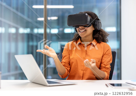 Smiling woman in orange shirt exploring virtual reality with VR headset, interacting with laptop in modern office. Concept of innovation, technology, and immersive experiences. Smiling woman in orange shirt exploring virtual reality with VR headset, interacting with laptop in modern office. Concept of innovation, technology, and immersive experiences. 122632956