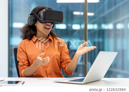 Young African American businesswoman using virtual reality headset while interacting with laptop in modern office. Emphasis on innovation, technology, and professional experience. Young African American businesswoman using virtual reality headset while interacting with laptop in modern office. Emphasis on innovation, technology, and professional experience. 122632984
