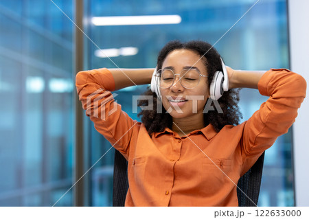Young African American businesswoman relaxing in modern office, leaning back in chair, eyes closed, wearing headphones and orange shirt. Scene conveys calm, focus and relief from work stress. 122633000