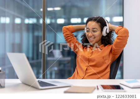 Smiling woman in orange shirt and headphones sitting at desk with laptop. Relaxed and happy during work break. Bright modern office space, casual and comfortable atmosphere. Smiling woman in orange shirt and headphones sitting at desk with laptop. Relaxed and happy during work break. Bright modern office space, casual and comfortable atmosphere. 122633001