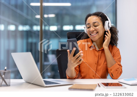 Smiling woman in orange shirt uses smartphone and headphones at office desk with laptop and notebook. Relaxed work vibe and enjoyment of digital music with phone. 122633002