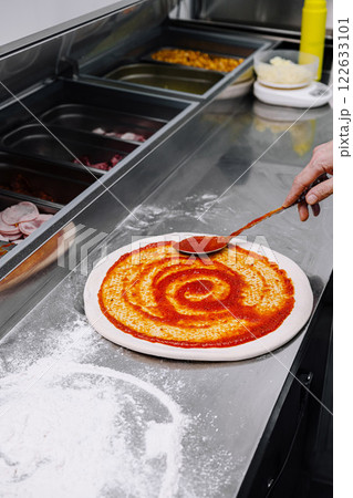 Preparing fresh pizza sauce in a busy kitchen with a hand spreading sauce on dough 122633101
