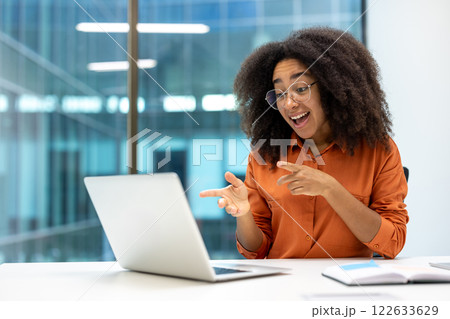 Young African American office worker excitedly pointing during online meeting. Businesswoman using laptop in office. Vibrant expression reflects engagement, communication, success, technology use. 122633629
