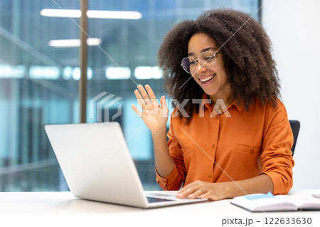 Smiling woman in orange shirt waves during online meeting, engaging in communication through laptop. Bright office setting conveys positive work-from-home environment and internet connectivity. 122633630